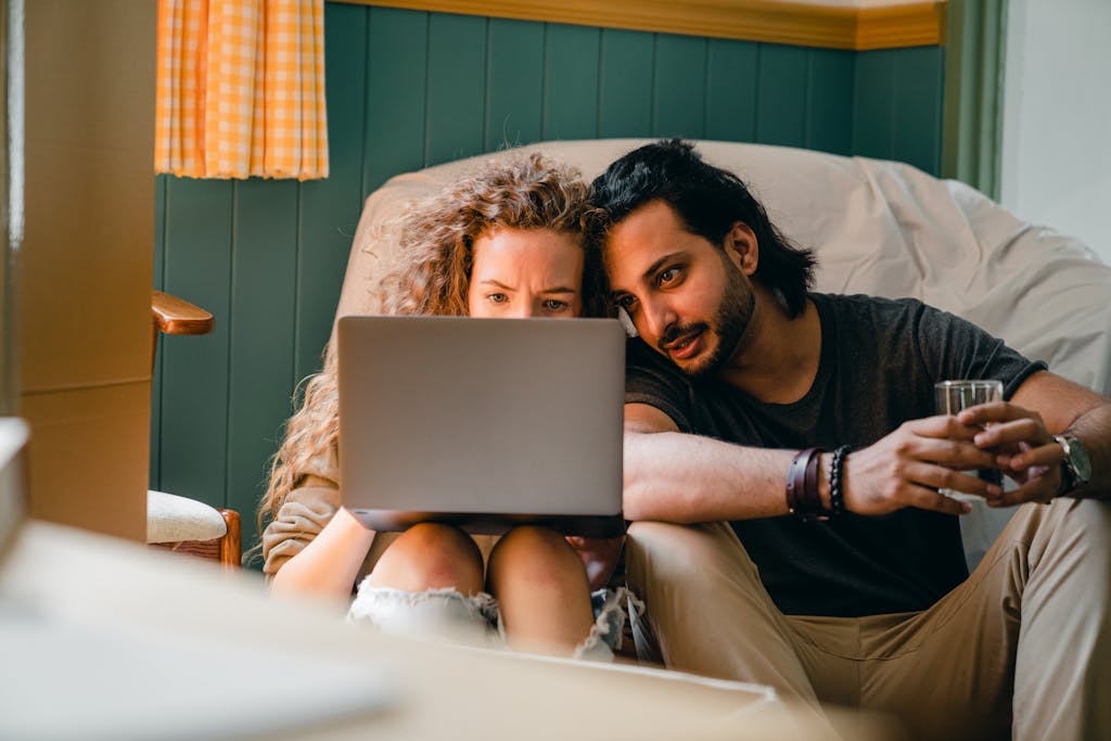 Focused young diverse couple in casual clothes sitting on floor against chair under white cover and browsing netbook