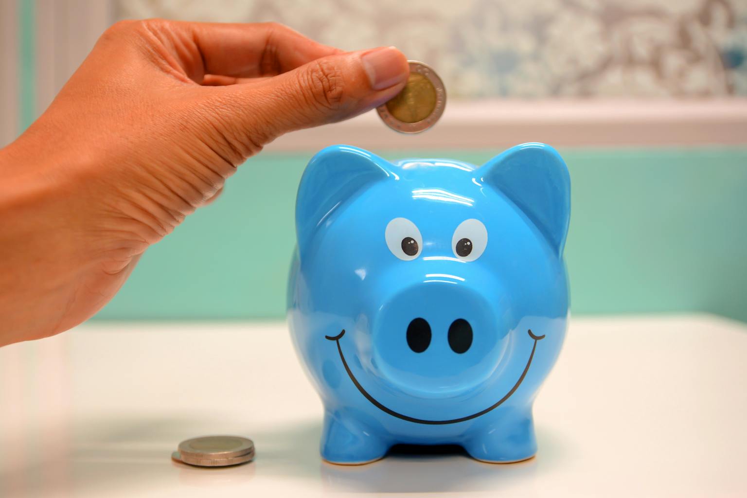 Une main insérant une pièce dans une tirelire bleu souriante en forme de cochon représentant la sécurisation de son épargne dans un livret